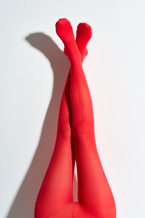 Long slender legs of young woman in red tights raised up over white wall background.