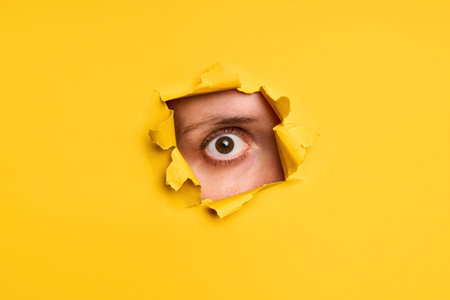 Close-up of human eye looking through torn yellow paper to camera