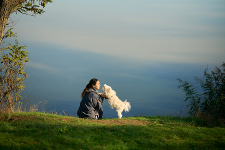 Woman kiss and hug her cute white dog sitting on grass over scenic mountain background. Owner and pet enjoying walking and time together. Love conceptの写真素材
