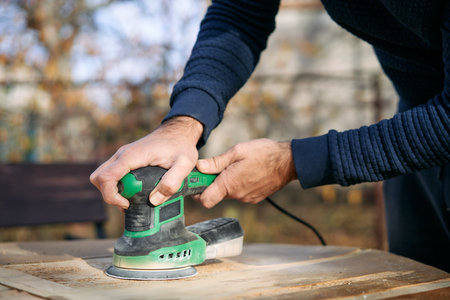 Close up of hands holding orbital sander while polishing wooden surface during renovation outdoorsの写真素材