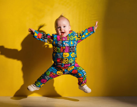 Adorable toddler child in a bright patterned jumpsuit jumping high against a yellow wall with shadow. Generative AIの素材