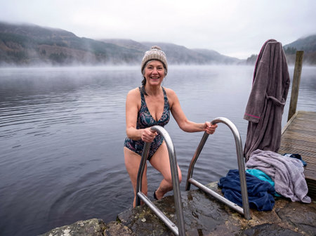 Mature woman exiting cold lake via wooden ladder after winter swimming session in foggy weather. Generative AIの素材