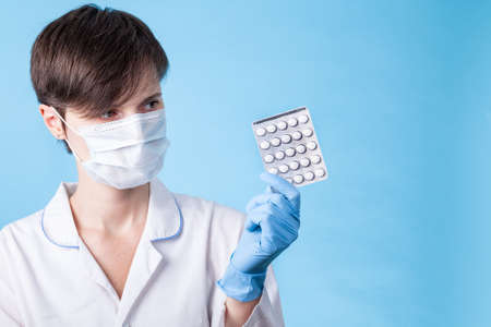 A young attractive female scientist in a mask holds a package of pills with gloved fingers on a blue...