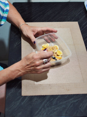 woman's hands eating sliced ââââbanana with Peruvian maca oats in a clear pot with spoon on a dark marble table and table protectorの写真素材