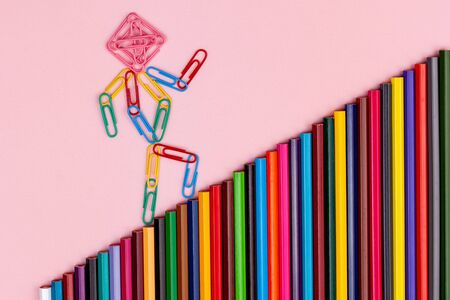 man from multi-colored paper clips goes up the stairs from multi-colored pencils isolated on a pink backgroundの写真素材