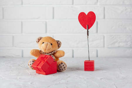 on a white tectonic background a teddy bear with a red gift box for the ring, on the right a red heart on the mount, in the background a white brick wallの写真素材