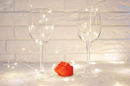 two empty wine glasses stand on a light textured surface, between glasses a red box, around the glasses there is a glowing garland, in the background a white brick wallの写真素材