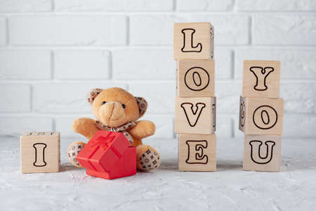 Against the white brick wall a small teddy bear with a red box and wooden cubes with letters, laid out the phrase I love youの写真素材