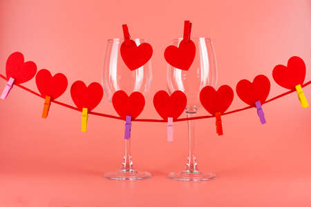 in the foreground is a garland on a red ribbon with red hearts, in the background two wine glasses on a pink backgroundの写真素材