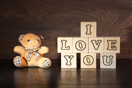 on a dark wooden background of wooden cubes with letters laid out phrase I love you, to the left of the cubes sits a small teddy bear, used as a background or postcardの写真素材
