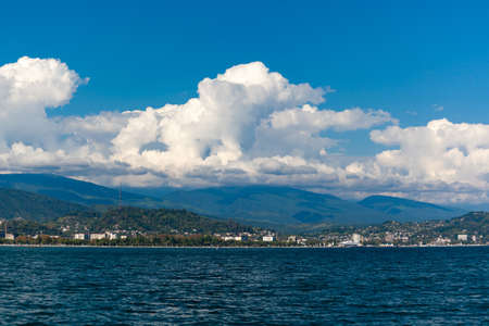 Sea and mountains against the blue sky with white clouds, at the bottom of the mountains are buildingsの写真素材