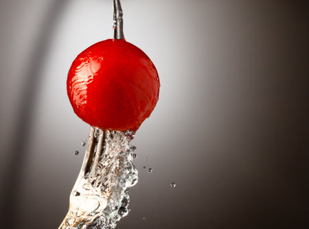 red cherry tomato on a silver fork. A jet of water, splashes of water. Black white gradient background. Place under the textの写真素材