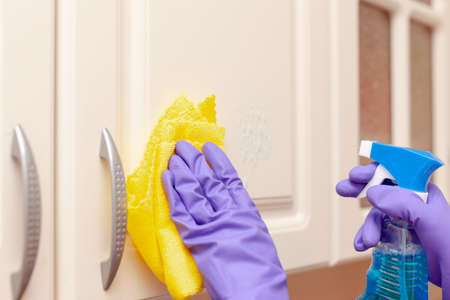 female hand in a purple rubber glove wipes the beige kitchen cabinet door. Yellow rag in hand