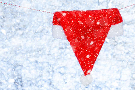 A red Christmas cap hangs on a rope. Against the backdrop of a blurred shiny white-blue background. Concept. Template for design. Copy spaceの写真素材