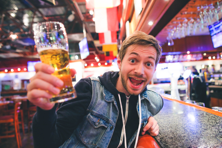 Portrait of emotional bearded hipster man with glass of beer cheering sitting at sports barの写真素材