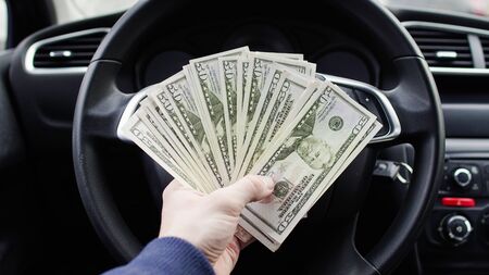 A dollar bills in the male hand on the background of the steering wheel in the car. Concept of...