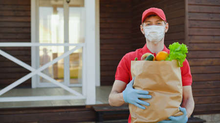 Portrait of deliverer in red uniform wearing safety mask holding a box of grocery food. Delivery service in quarantineの写真素材