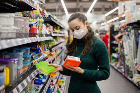Woman wearing protective face mask chooses new plastic containers for storing food in supermarket....