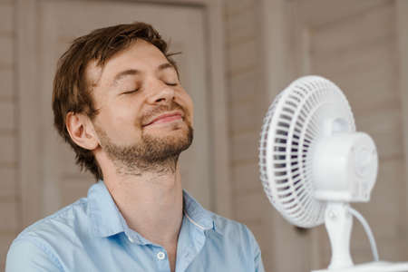 Sweaty man enjoying air flow from fan in office. Businessman refreshing in front of air electric ventilator.の写真素材