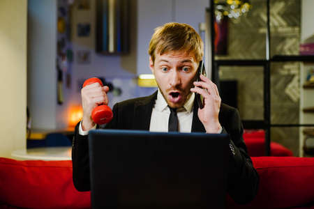 Overworked businessman in suit speaking on smartphone, working on laptop, reading writings in laptop and training with dumbbell at the same time. Multitasking. Busy life conceptの写真素材