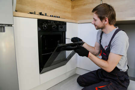 Young man worker in uniform repairing electric oven in kitchen.の写真素材