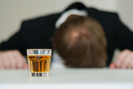 Drunk stressed businessman sleeping at office desk. Alcohol addiction. Shot of whisky in the foreground.の写真素材
