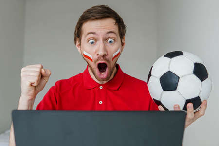 Young excited man fan support Poland national football team hold in hand soccer ball watch tv live stream on laptop.の写真素材