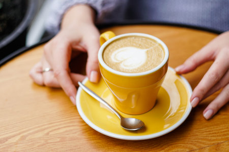 Woman holding hot yellow cup of latte coffee on a wooden table.の写真素材