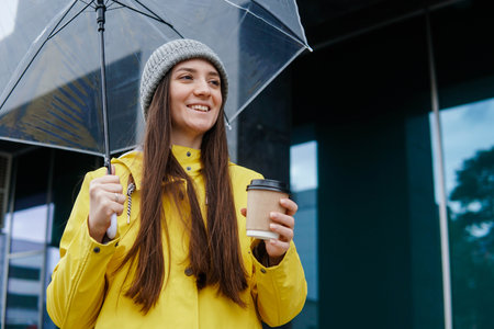 Portrait of pretty young girl in yellow raincoat holding umbrella and cup of coffee to go in the street.の写真素材