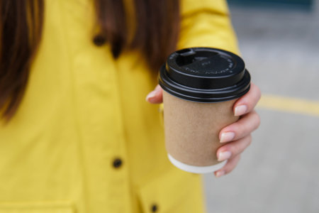 Close up of young woman in yellow raincoat holding paper cup of coffee takeaway in the street.の写真素材