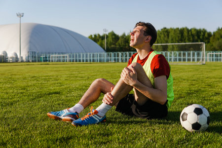 Young man football player suffering from leg injury sitting on the grass at soccer stadium.の写真素材