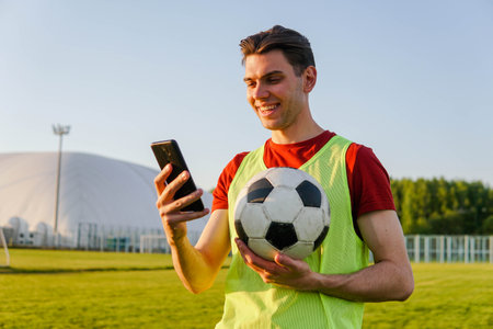 Portrait of young smiling man football player holding soccer ball and using smartphone