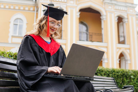 Happy smiling female graduate student in black academic gown with laptop over university campus backgroundの写真素材