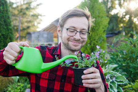 Young funny man florist in glasses holds a green watering can and watering plant outdoorsの写真素材