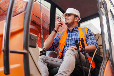 Young man loader worker or a forklift driver in a container warehouse wearing a white helmet is talking on the walkie-talkie or radio phone.の写真素材