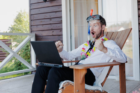 Young happy businessman in hawaiian necklace and snorkeling mask sitting in wooden chair and using laptop.の写真素材