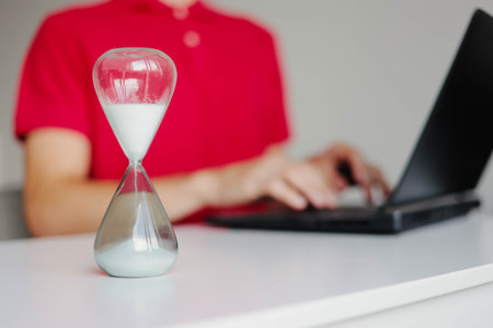 Hourglass with flowing sand on wooden table. Man typing on a laptop keyboard indoors. Concept of...
