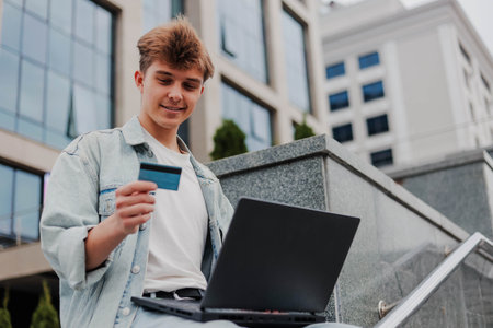 Happy young man using laptop and credit card for online shopping outdoors on a background of corporate building.の写真素材