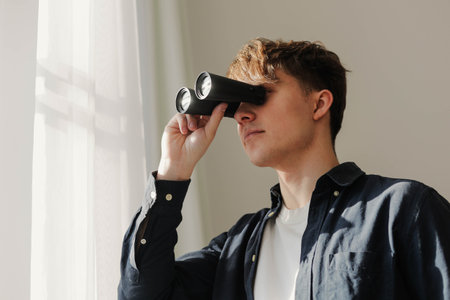 Young man watching through binoculars in front of a window.の写真素材