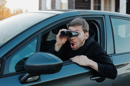 Young shocked man watching through binoculars sitting in car. Man spying or investigating.の写真素材