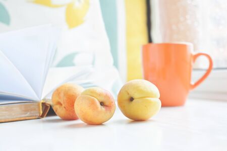 Three apricots on a window sill against the background of book, pillows and cup on the window. Set for a cozy summer reading.の写真素材