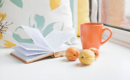 Three apricots on a window sill against the background of book, pillows and cup at the window on a rainy day. Set for a cozy summer reading.の写真素材