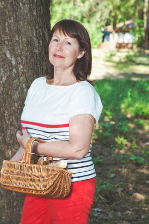 Cute elderly woman holding a wicker basket full of ceps and smiling at the camera next to a tree in a sunny warm forestの写真素材
