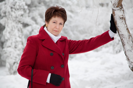 Attractive elderly woman in red coat standing near tree in winter snowy parkの写真素材