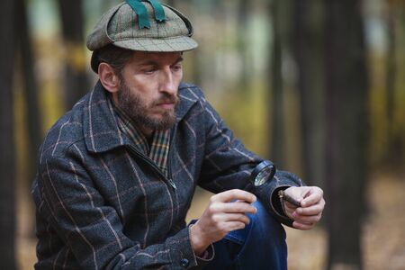 Imposing man detective with a beard wearing a cap and a plaid jacket considers through magnifying glass fragment of a tree in autumn forestの写真素材