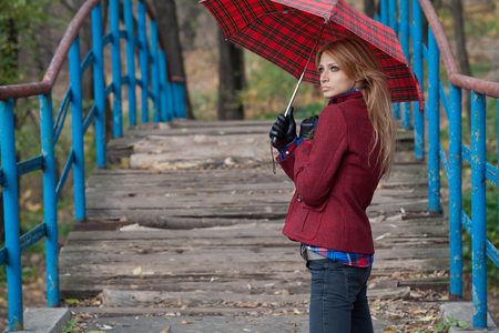 Glamorous pretty woman in jeans and burgundy jacket standing under umbrella on the bridgeの写真素材