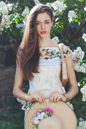Beautiful young girl model with straw hat posing near white blooming lilacs at sunny spring dayの写真素材
