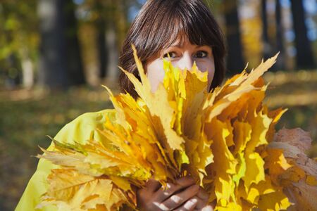 Elderly attractive woman hiding her face behind armful yellow autumn leaves in forest on sunny afternoonの写真素材