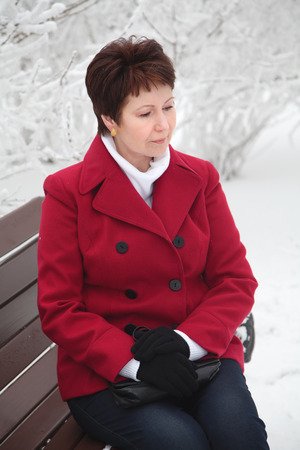 Attractive elderly woman in red coat sitting and resting on bench in winter snowy streetの写真素材