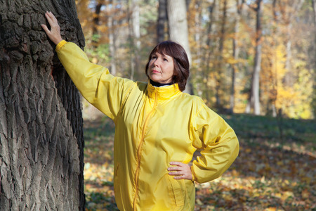 Elderly woman walks in autumn forest on a sunny day and enjoying the natural energy from the trunk of a large old treeの写真素材